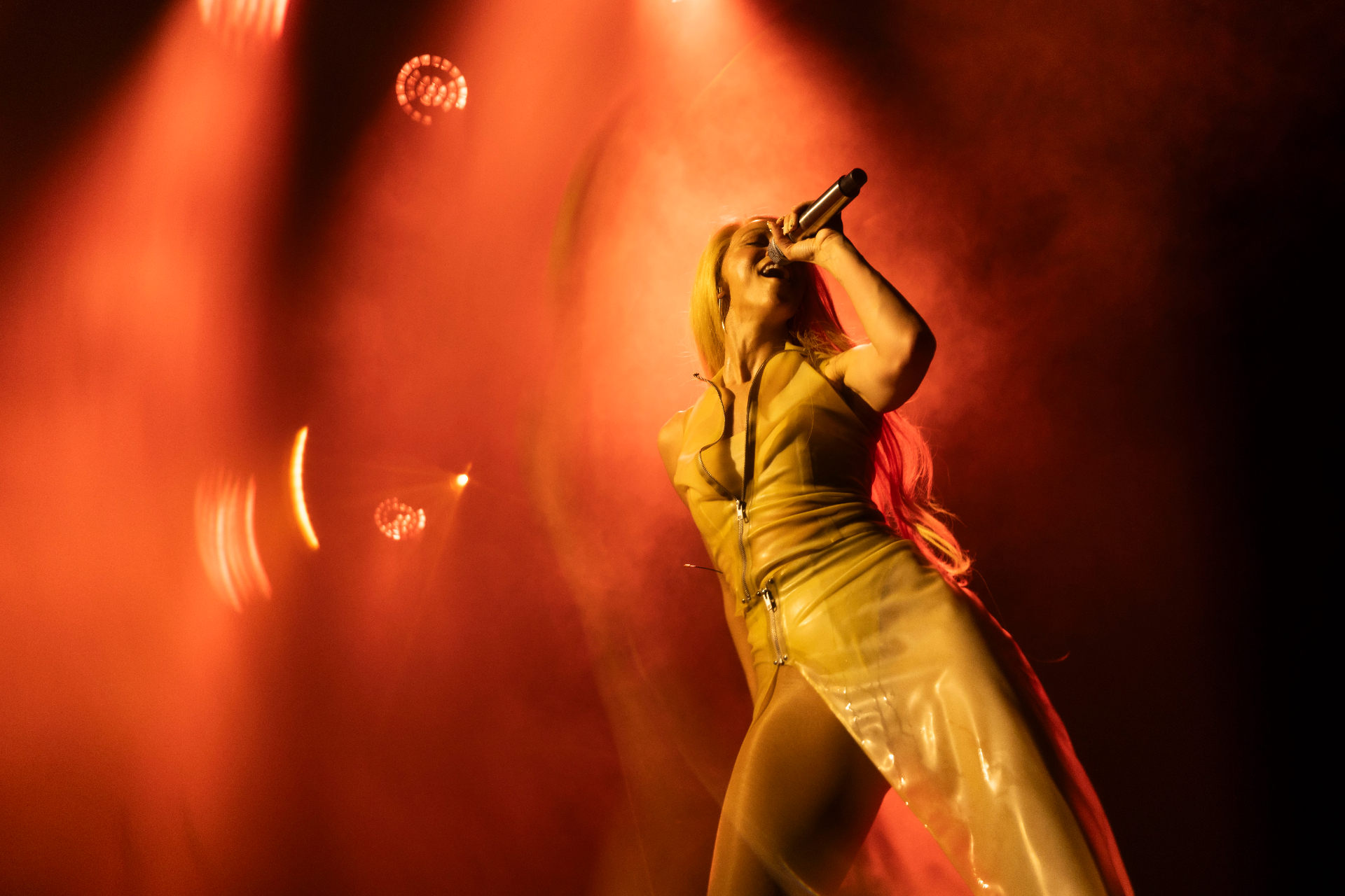 Agnes singing into a microphone with head tilted back, wearing a glossy yellow latex dress, lit by deep red and orange stage light