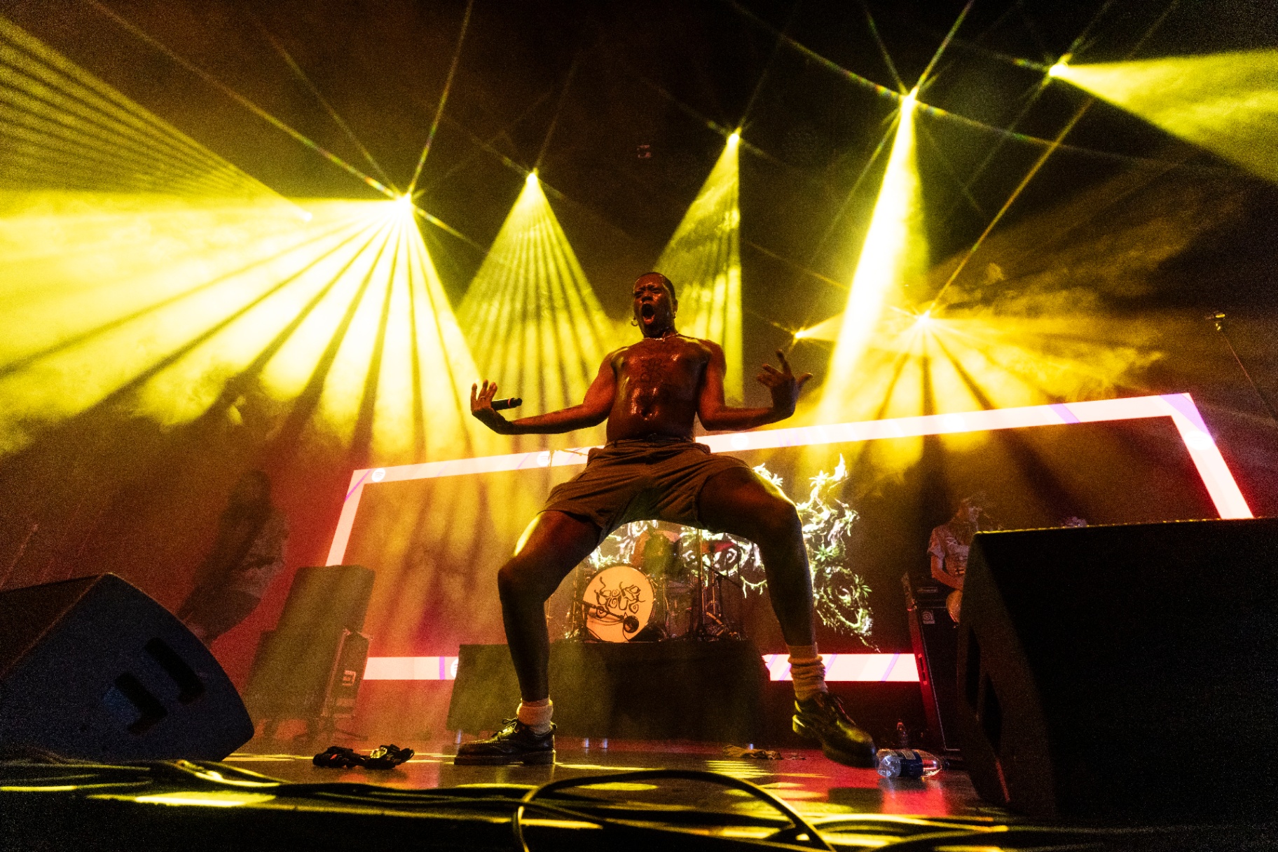 Boko Yout shirtless on stage with arms raised and mouth open mid-shout, framed by yellow laser fans and an LED rectangle behind him