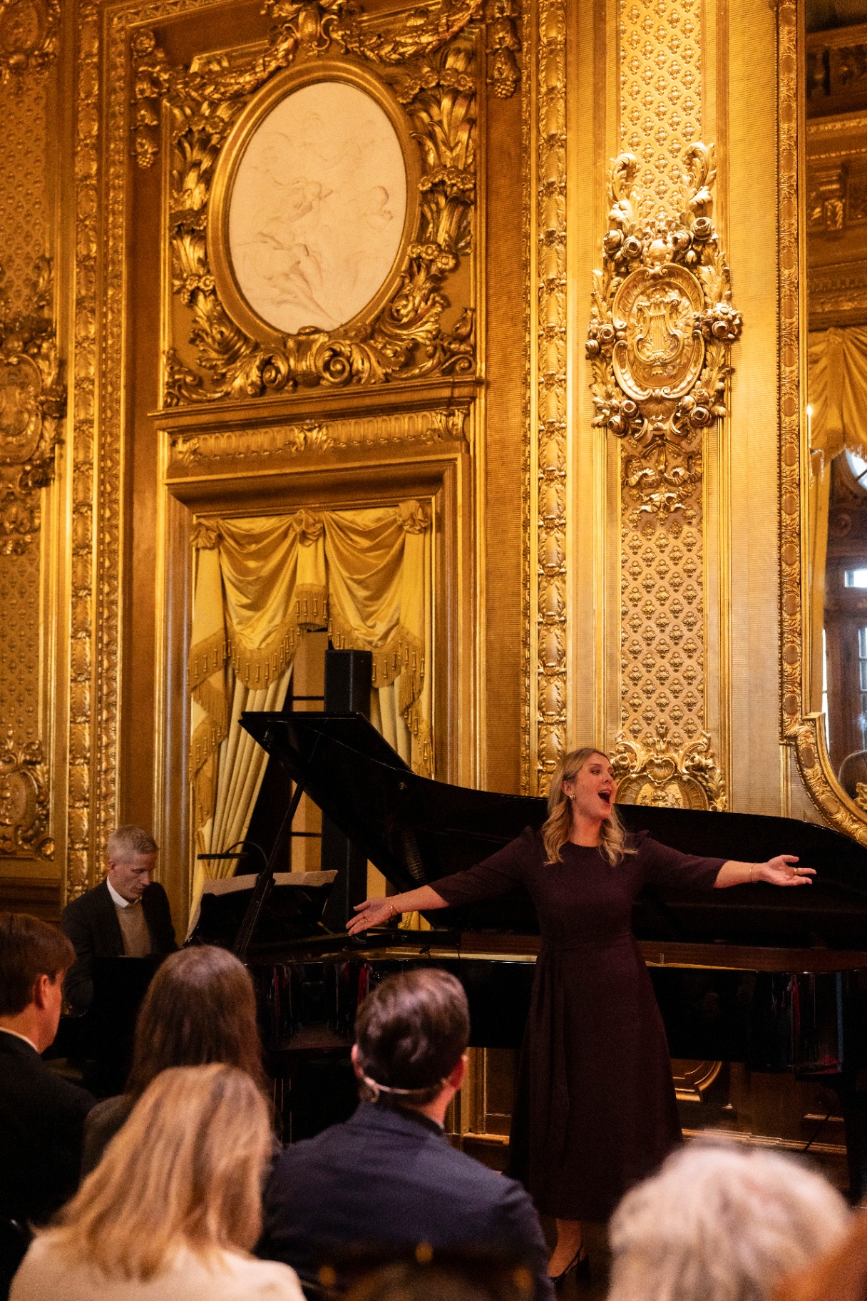 Christina Nilsson singing with arms outstretched in front of a grand piano, framed by gilded ornament and a chamber portrait