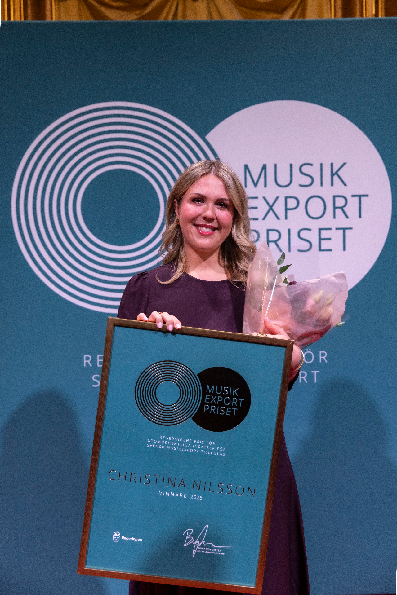 Christina Nilsson smiling, holding a framed Music Export Prize diploma and a bouquet of pink flowers