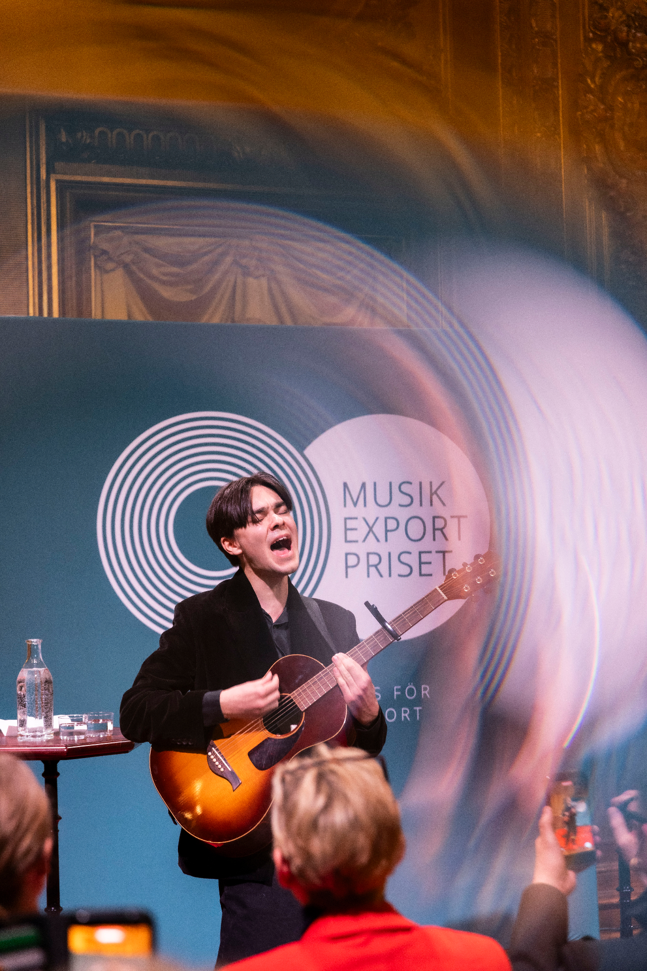 Isak Benjamin singing into the room with eyes closed, playing an acoustic guitar