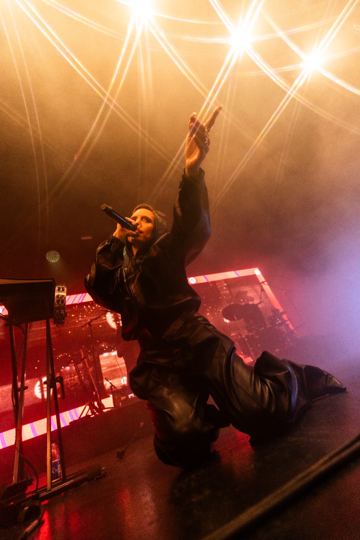 Lykke Li kneeling on stage in a black leather outfit, microphone close to her mouth and one arm raised toward the rig of white spotlights above