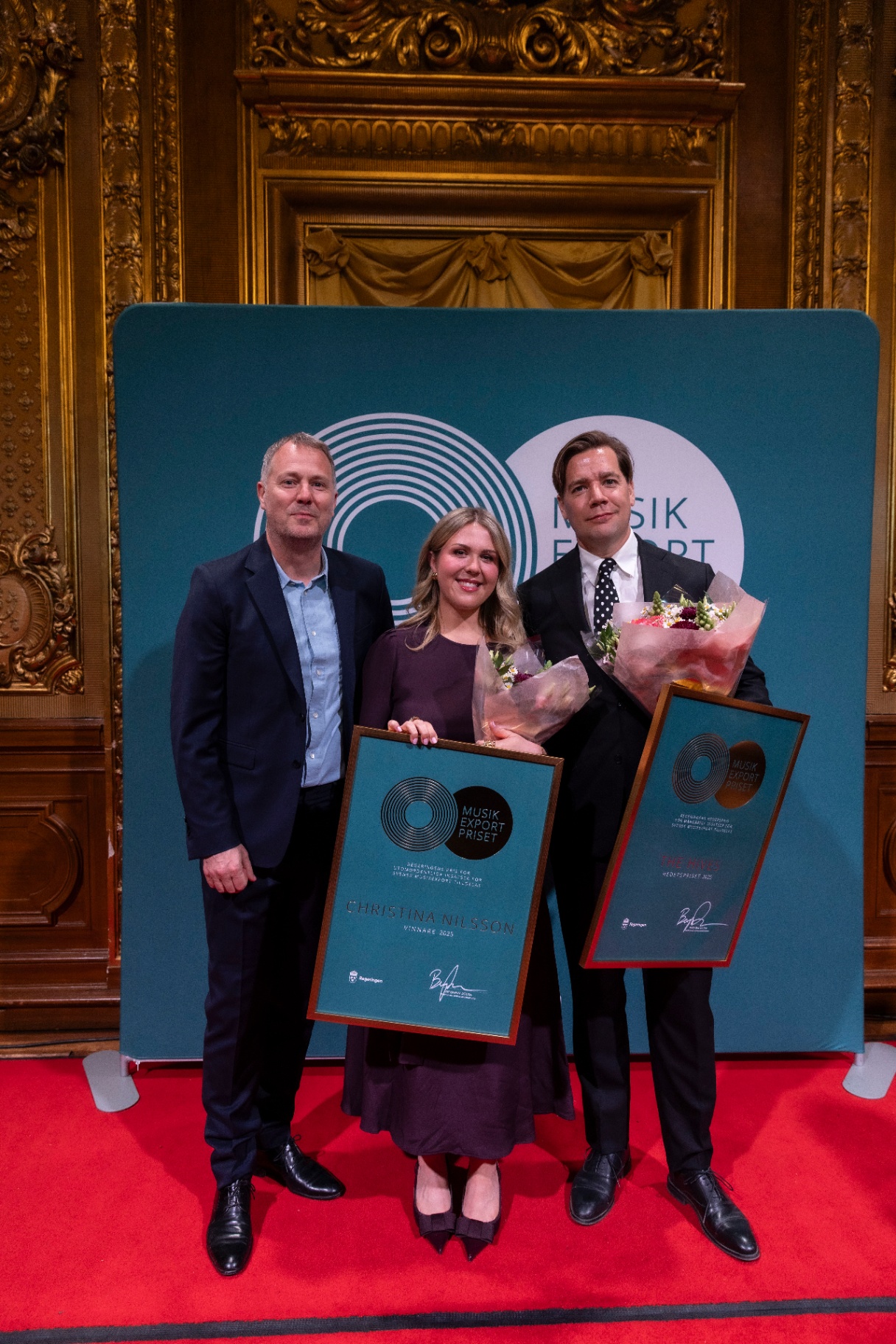 Johan Seidefors, Christina Nilsson and Pelle Almqvist standing on a red carpet, holding their framed diplomas and bouquets