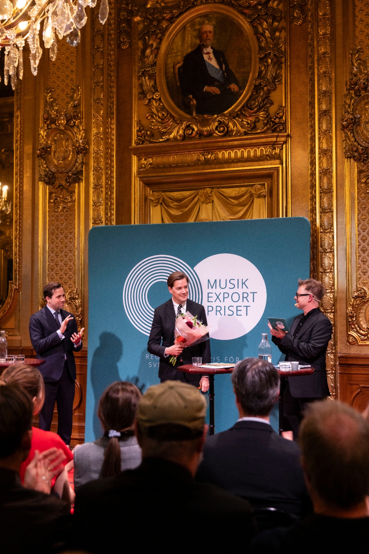 Pelle Almqvist on stage holding a bouquet of flowers, flanked by two presenters applauding, beneath a portrait and gilded ornament