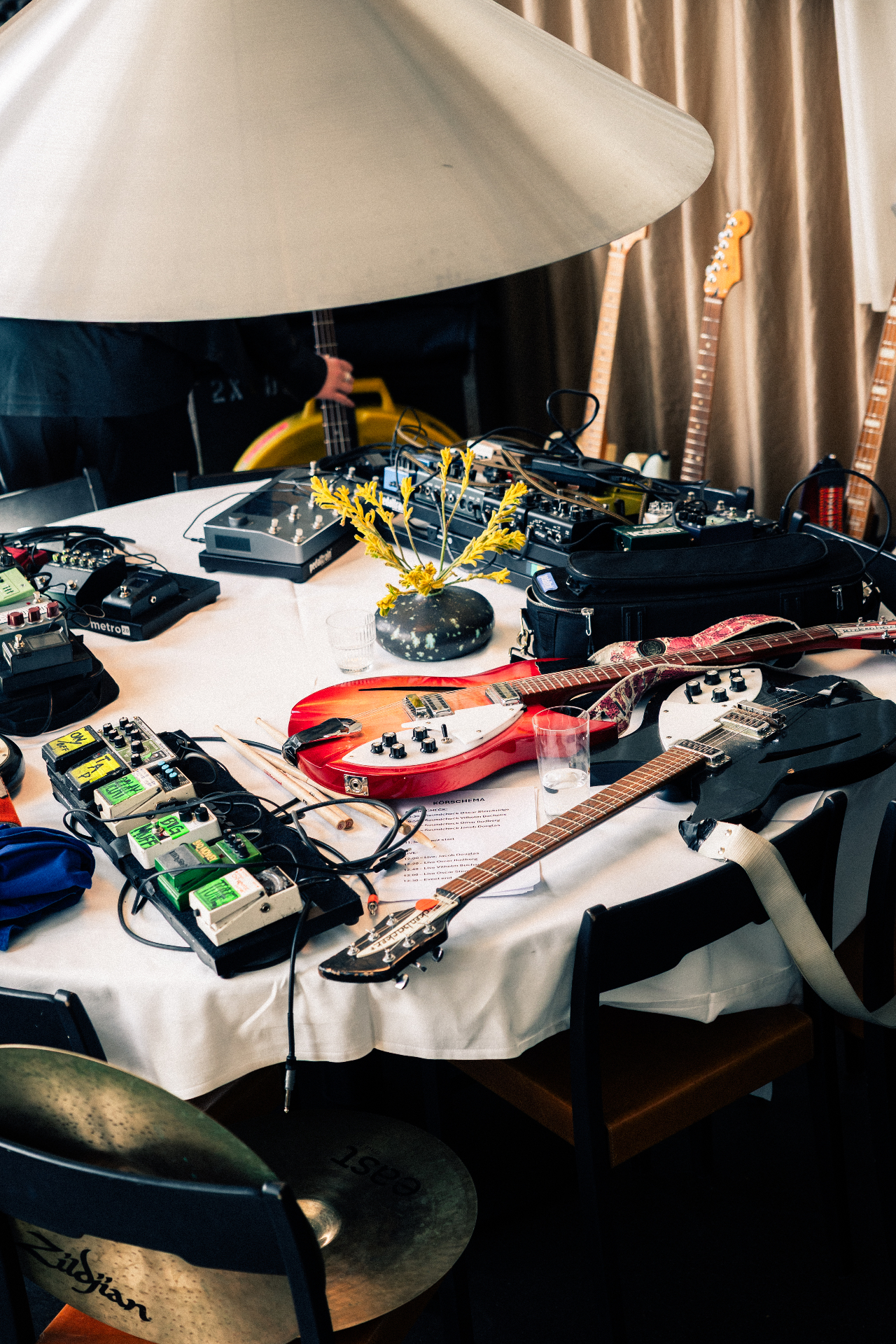 A round white-clothed table at Solen covered in guitar pedals, drumsticks, a red electric guitar and the printed run-of-show for the Amuse and TEN Music Group lunch, with two more guitars leaning against the wall behind.