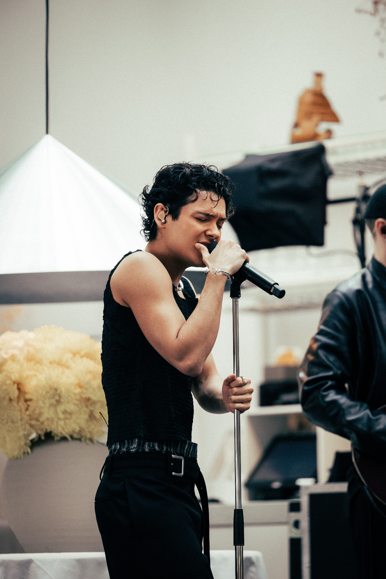A vocalist in a black tank top sings into a microphone on a stand at the Amuse and TEN Music Group lunch showcase at Solen, soft daylight and yellow flowers in the background.