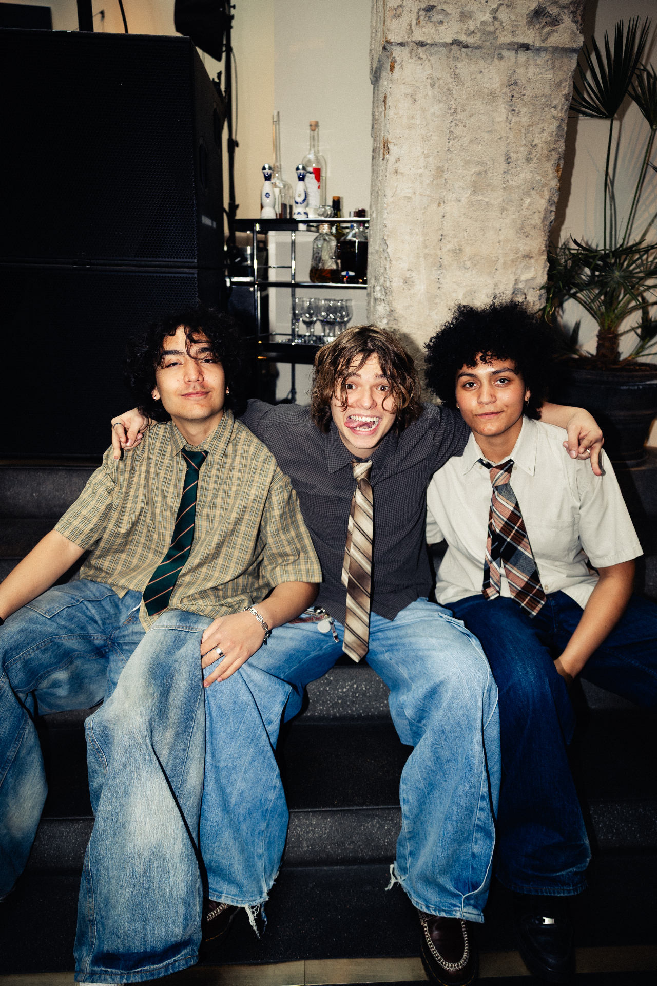 Three young artists in striped and plaid ties, button-down shirts and wide-leg jeans sit shoulder to shoulder on a step at Solen, smiling and laughing for the camera after the Amuse and TEN Music Group lunch showcase.
