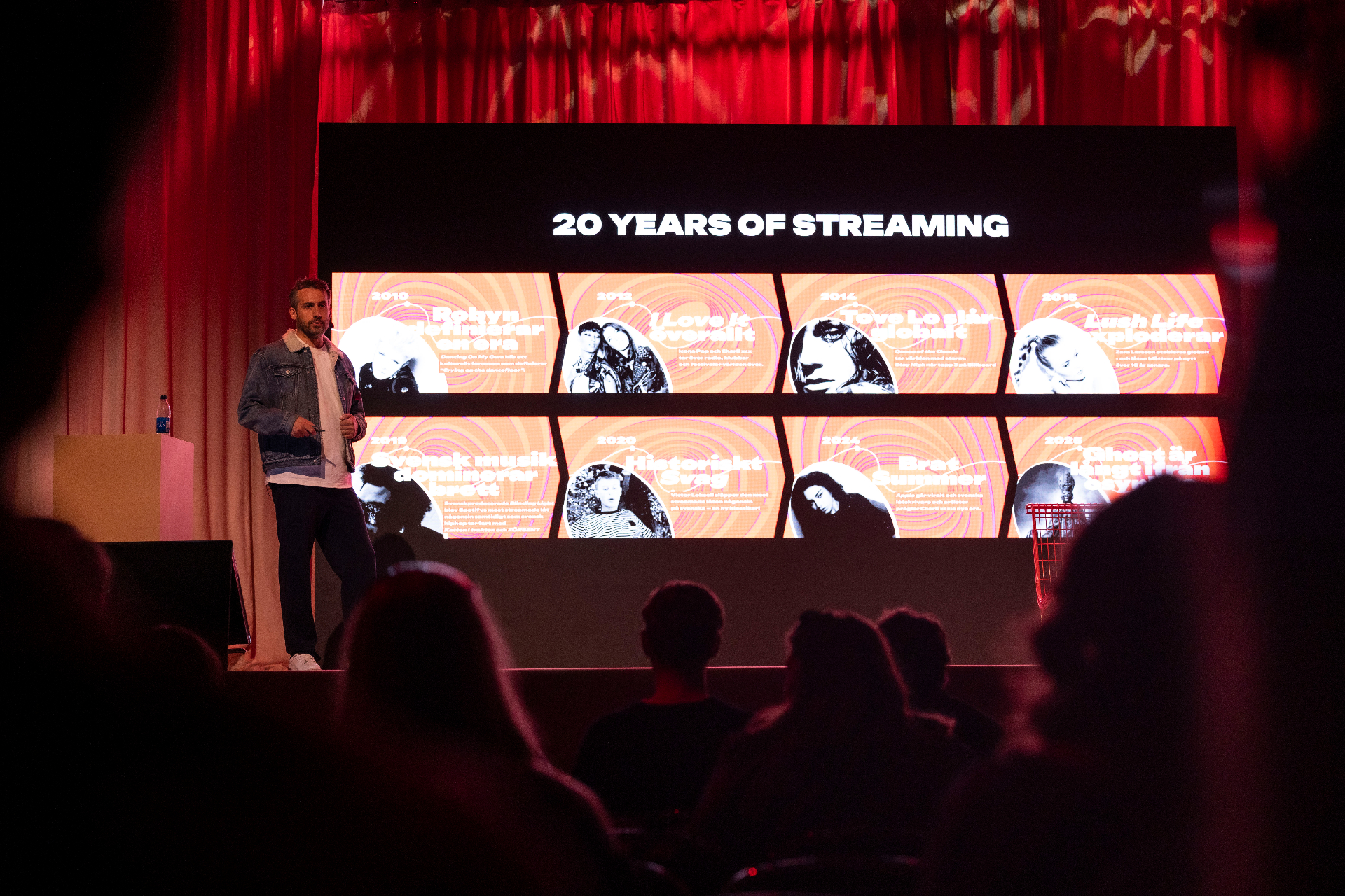 Andy Sloan-Vincent on stage at Fållan in front of a large screen reading "20 Years of Streaming", an eight-panel timeline of Swedish music moments from 2010 to 2025 displayed behind him.