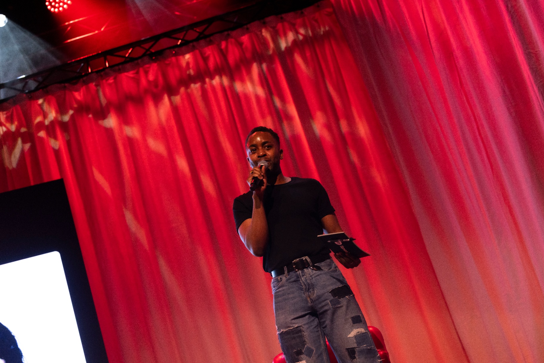A host in a black t-shirt holding a microphone on stage at Fållan, framed by a tall red curtain washed in red light.