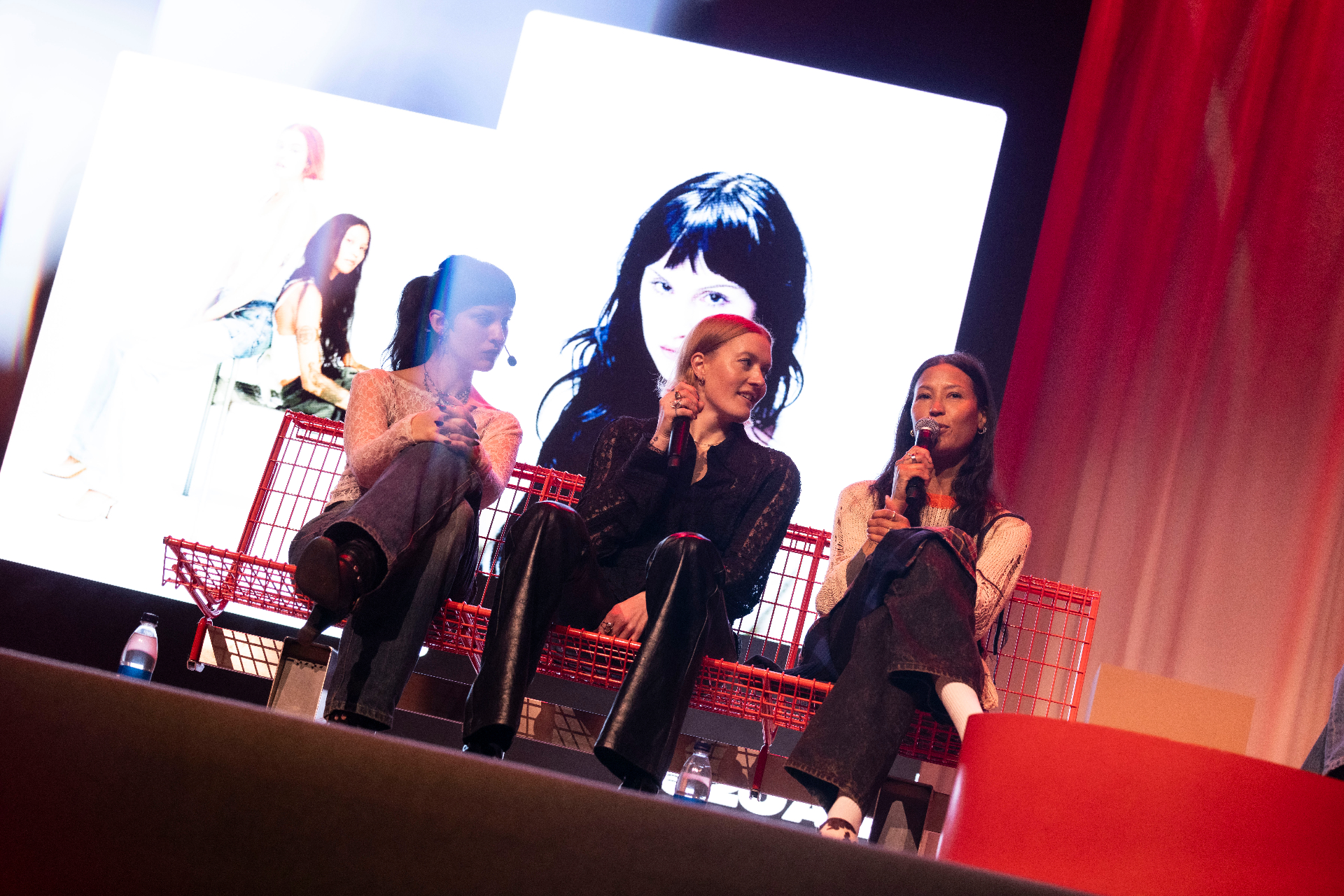 Olga Myko, Caroline Hjelt and Aino Jawo seated on a red wire bench at Fållan during the Artist to Artist conversation, large portraits of Olga Myko projected behind them.