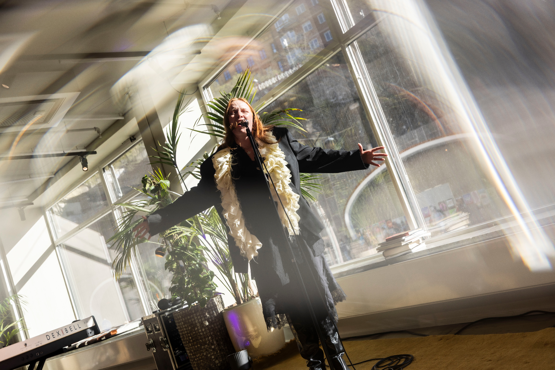 GERD performing live at AHouse, arms outstretched mid-song at the microphone in a black suit with a cream ruffled blouse, sunlight streaming through the tall windows behind her and palm leaves framing the shot.