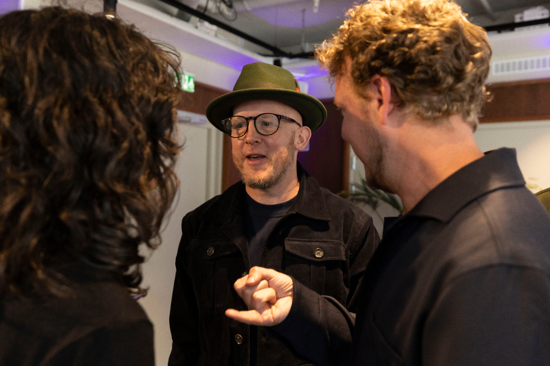 A guest in a green hat and corduroy jacket gesturing mid-sentence between two other attendees at the IFPI Industry Power Dinner.