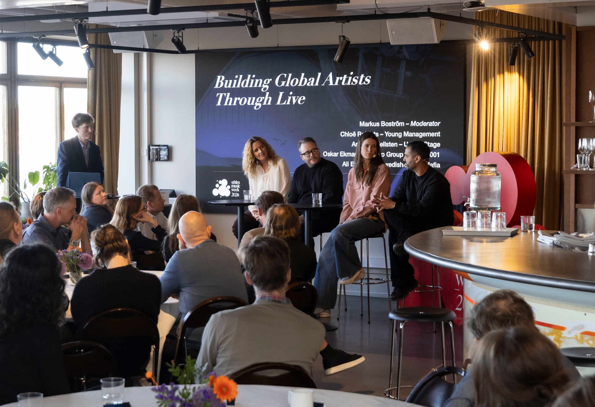 Building Global Artists Through Live panel on stage at AHouse, four panelists seated in front of a dark blue slide listing Markus Boström as moderator with Chloë Roberts, Anna Hellsten, Oskar Ekman and Ali Eskandari, audience watching from round tables in the foreground.