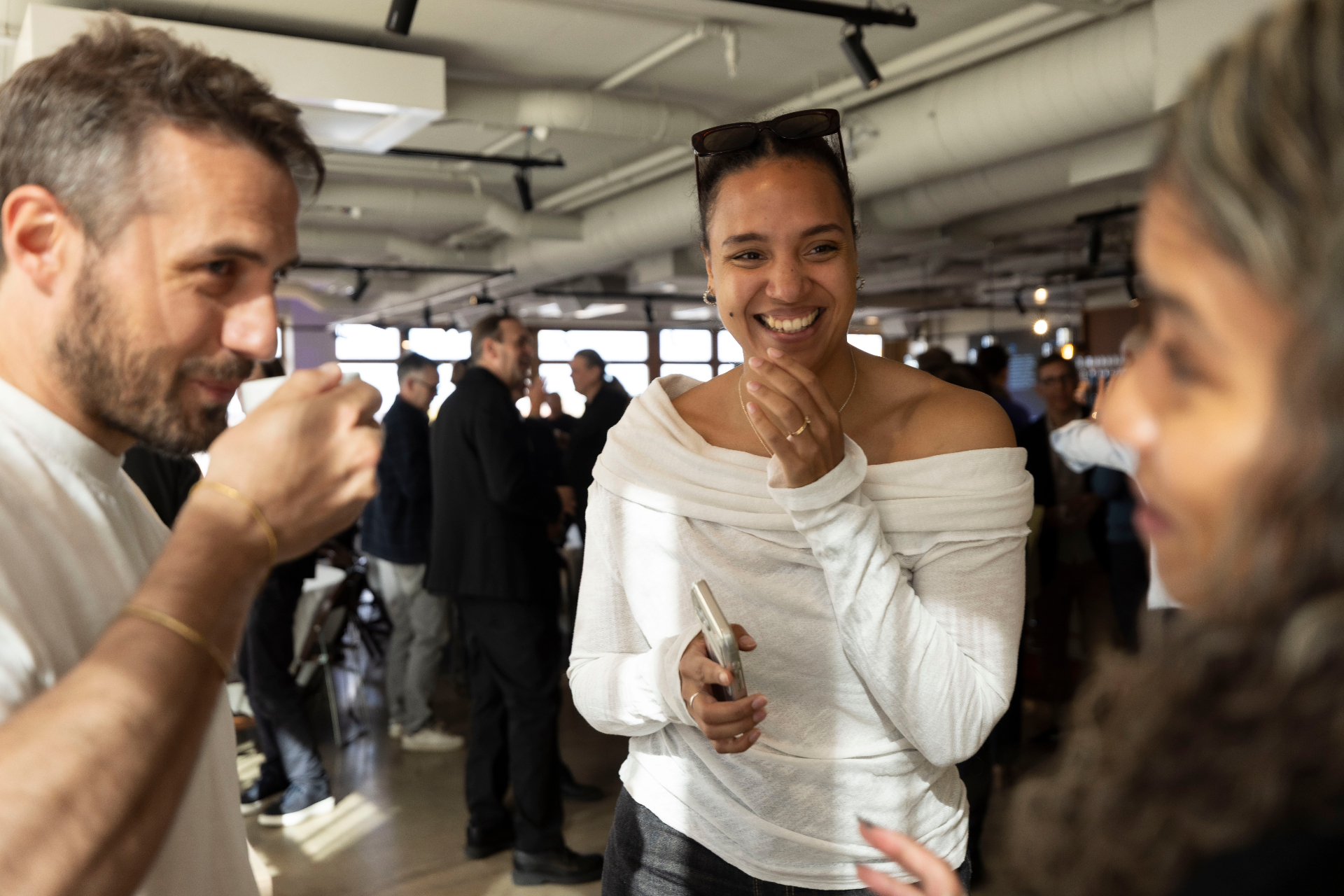 A smiling guest holding a phone in a white off-shoulder top, mid-laugh during the mingle at the IFPI Industry Power Dinner, with the room full of attendees behind her.