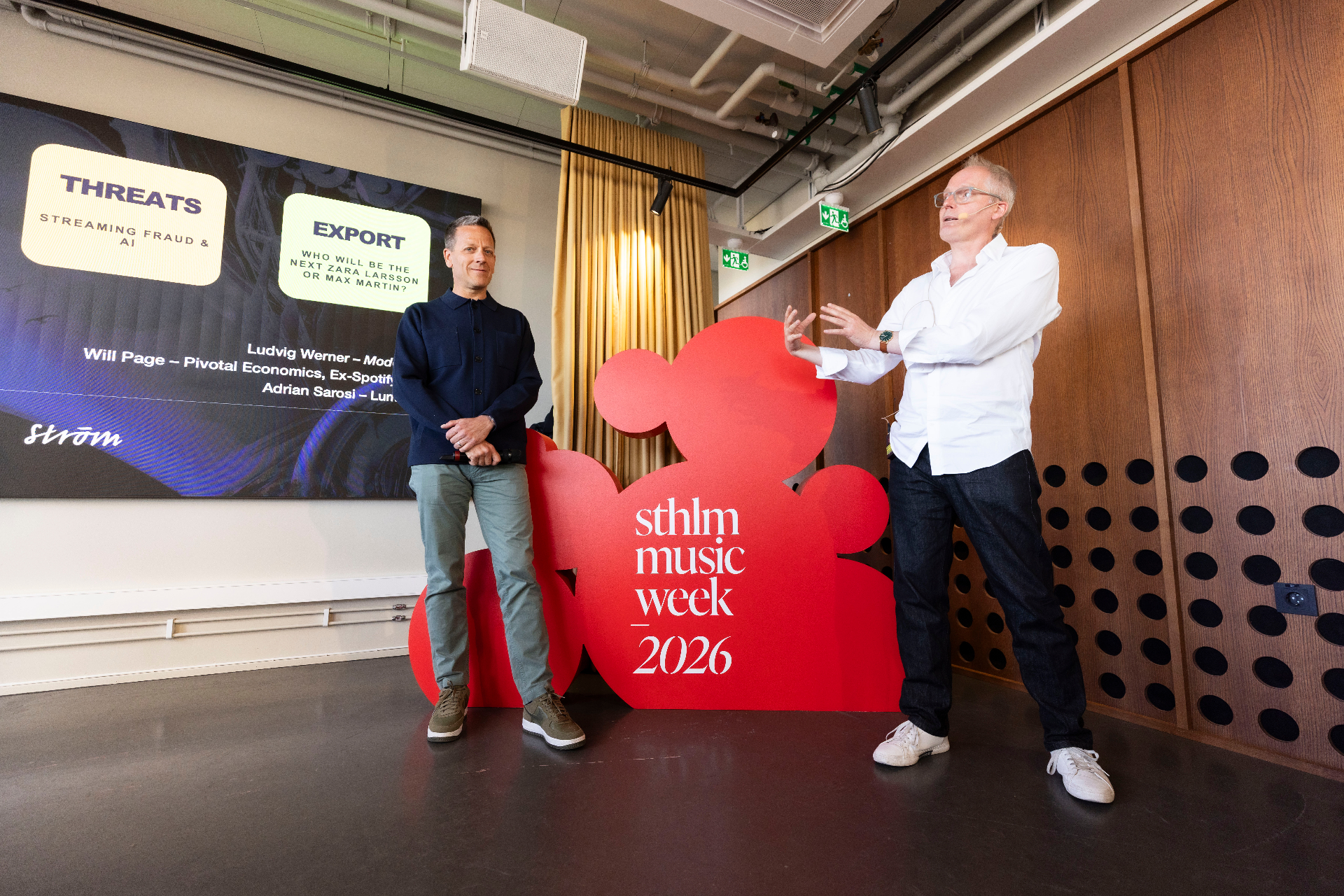 Two panelists standing on either side of the red sthlm music week 2026 sculpture, slide behind them showing Threats — Streaming Fraud & AI, and Export — Who will be the next Zara Larsson or Max Martin, with Ludvig Werner, Will Page and Adrian Sarosi listed.
