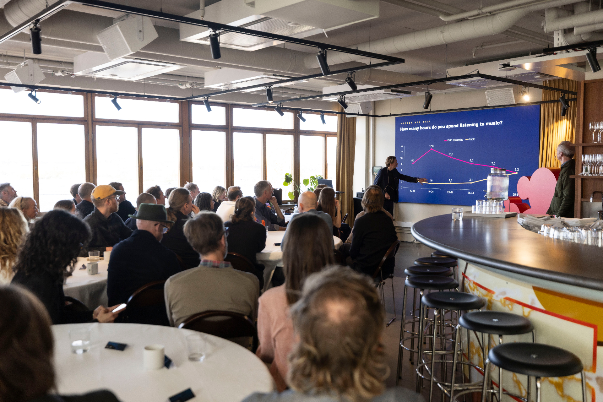 A speaker presenting the IFPI Global Music Report on a large blue screen showing a chart of paid streaming versus radio listening hours by age group, with the audience seated at round tables in the foreground.