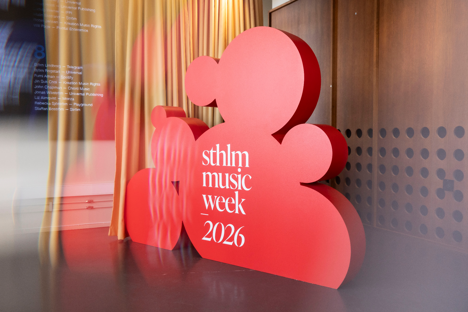 A large red Stockholm Music Week 2026 cloud-shaped sculpture standing in front of golden curtains at AHouse, with a printed guest list reflected in the glass behind it.