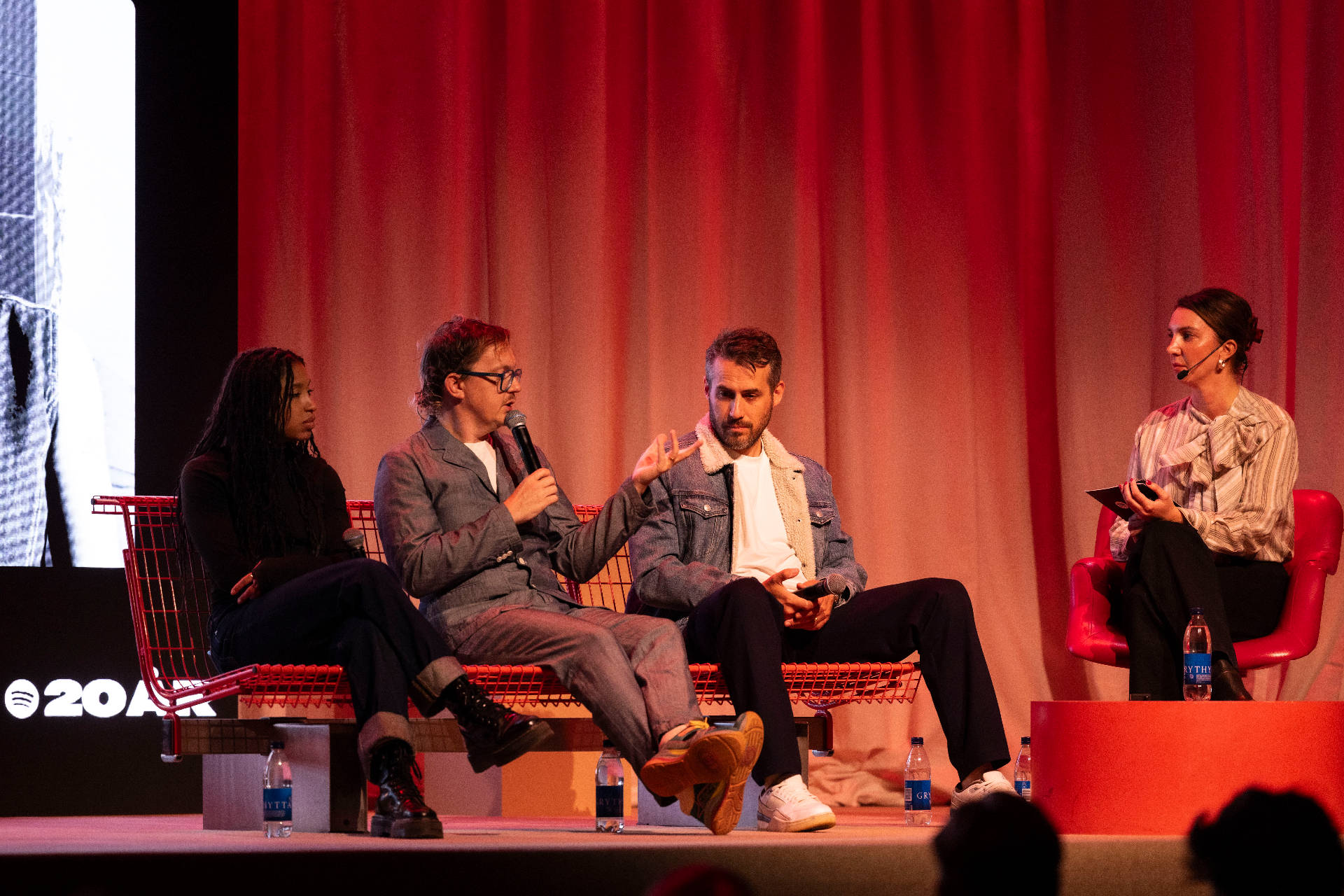 Panel on stage at Fållan in front of a deep red curtain, three guests on a red wire bench with a moderator on a red chair to the side.