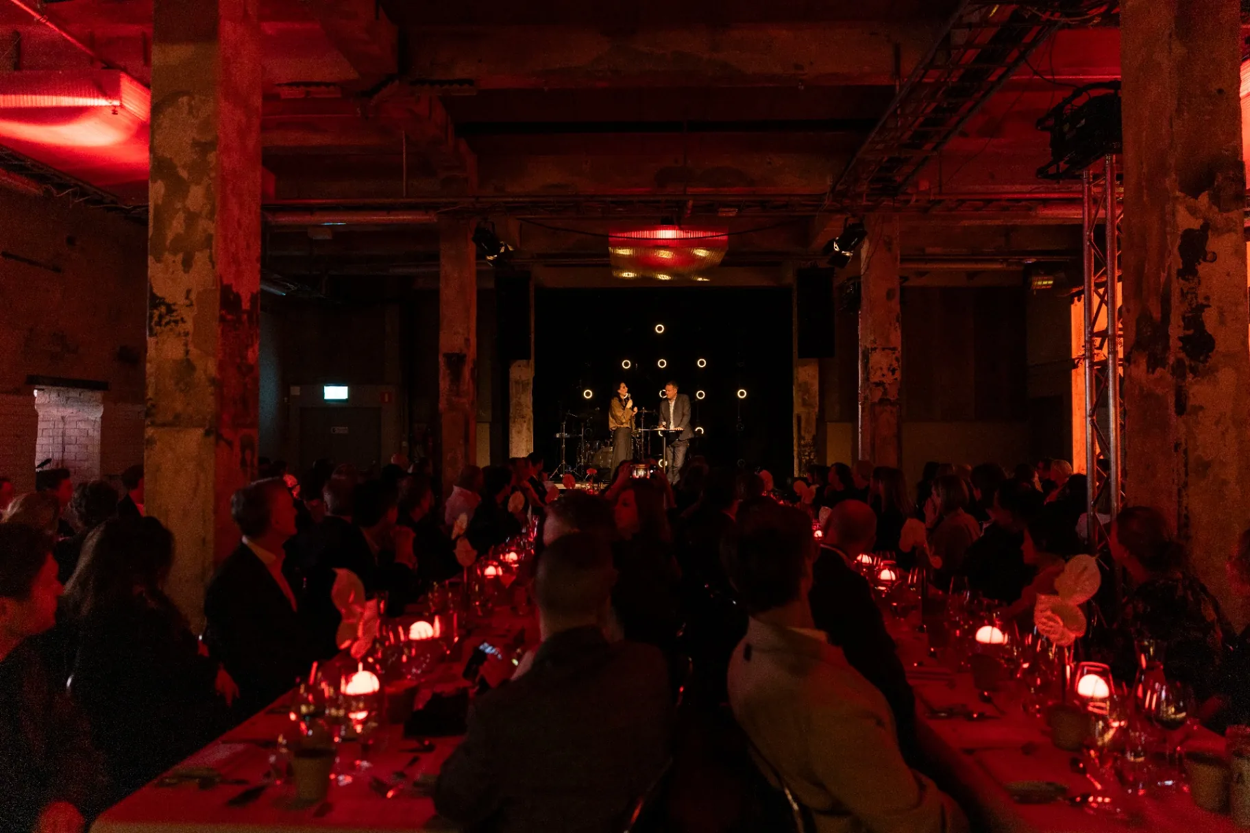 A long candlelit dinner table in a former industrial hall, guests facing a small stage at the far end